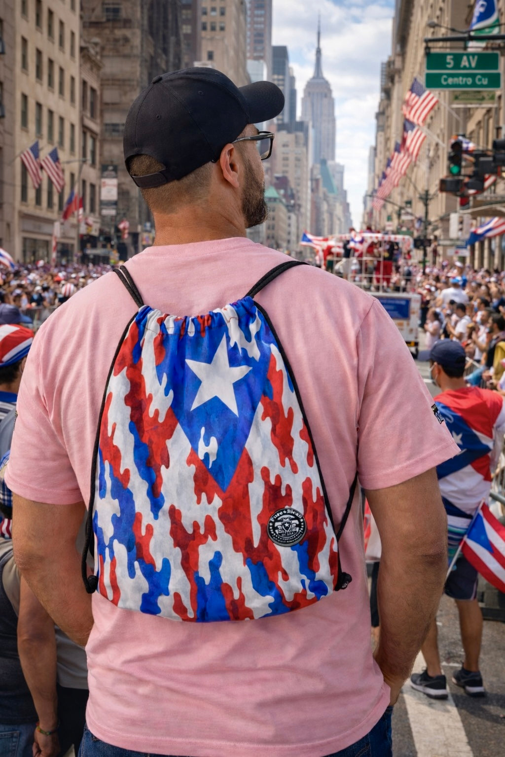 Pure⭐️Rican drawstring backpack worn by male model at Puerto Rican parade in New York featuring red white and blue camo flag design with crowd and city backdrop