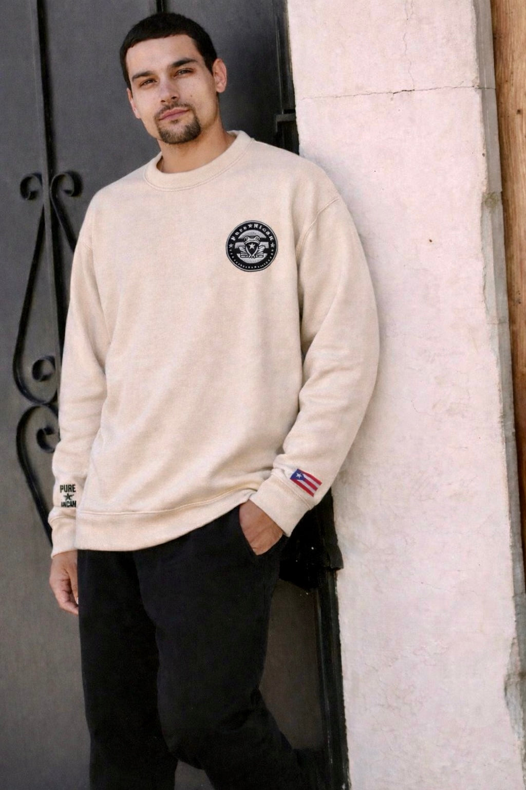 Man wearing a cream-colored sweatshirt with logos, standing against a textured wall.